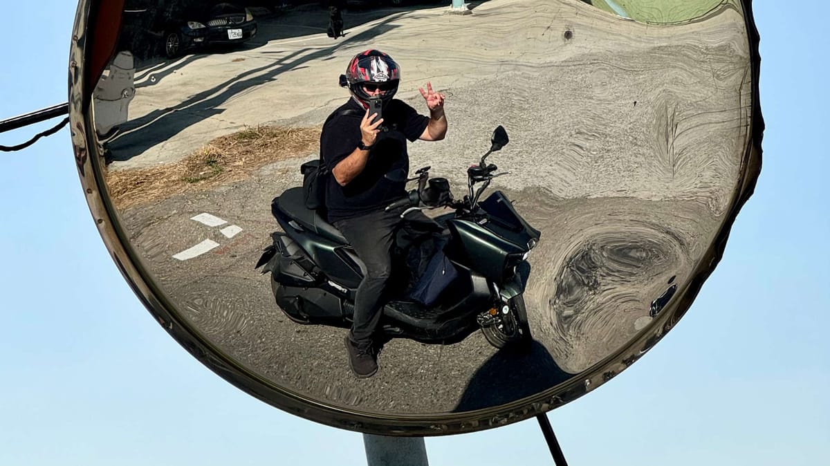 Distorted mirror reflection of a man riding a motorcycle in Taiwan, giving a peace sign.