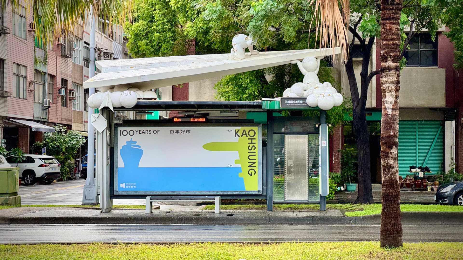 A bus stop. The roof is supported by blog-like figurines. A wide-format poster advertises Kaohsiung City’s 100th anniversary.