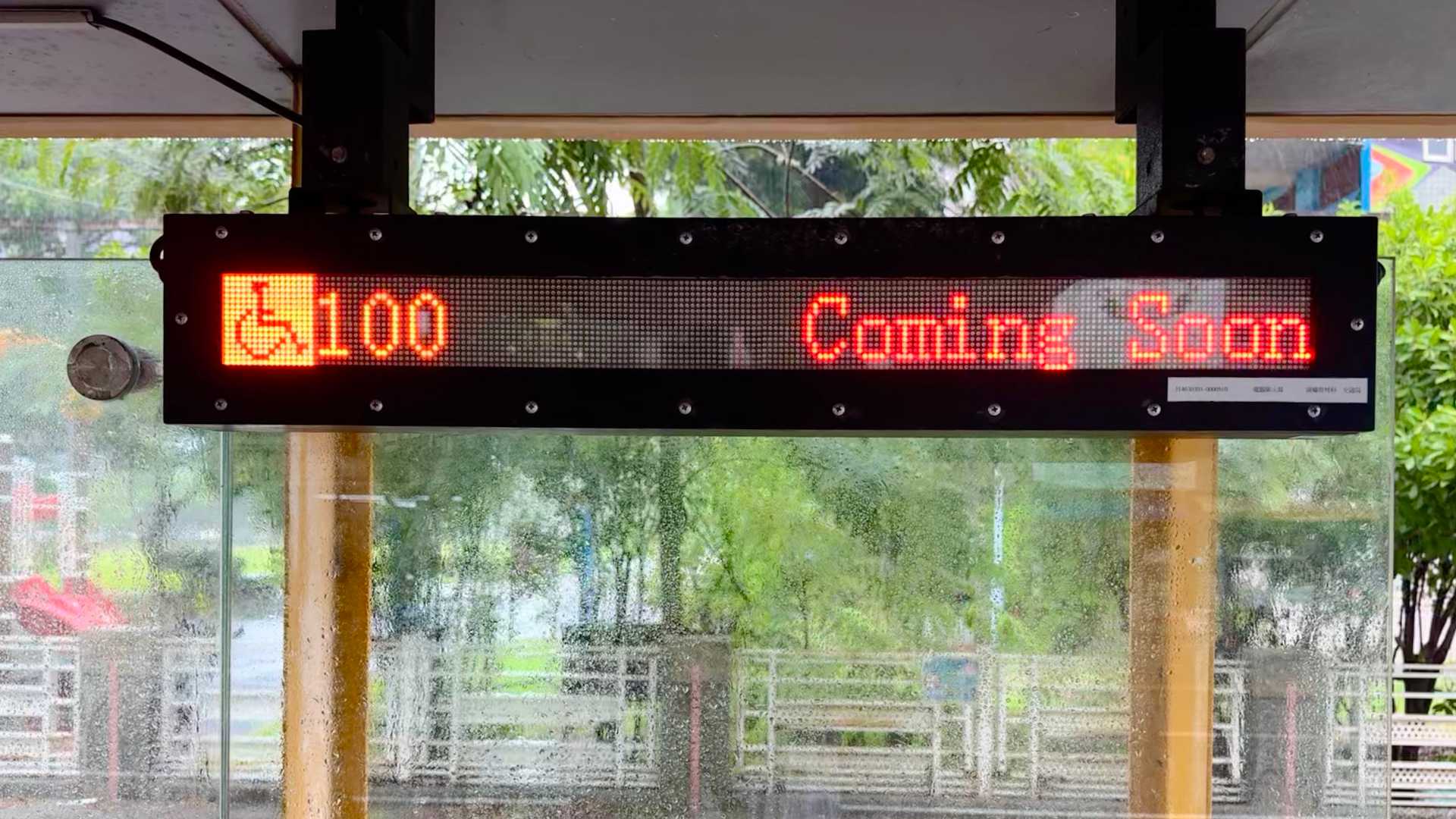 A dot-matrix sign at a bus stop, indicating bus number 100 is Coming Soon.