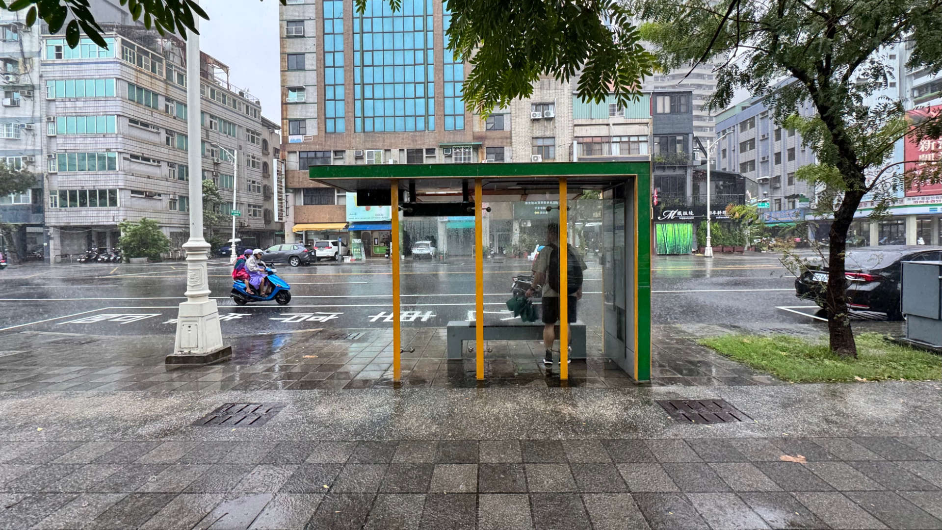 Person standing under a bus shelter in Kaohsiung, Taiwan, in the rain.