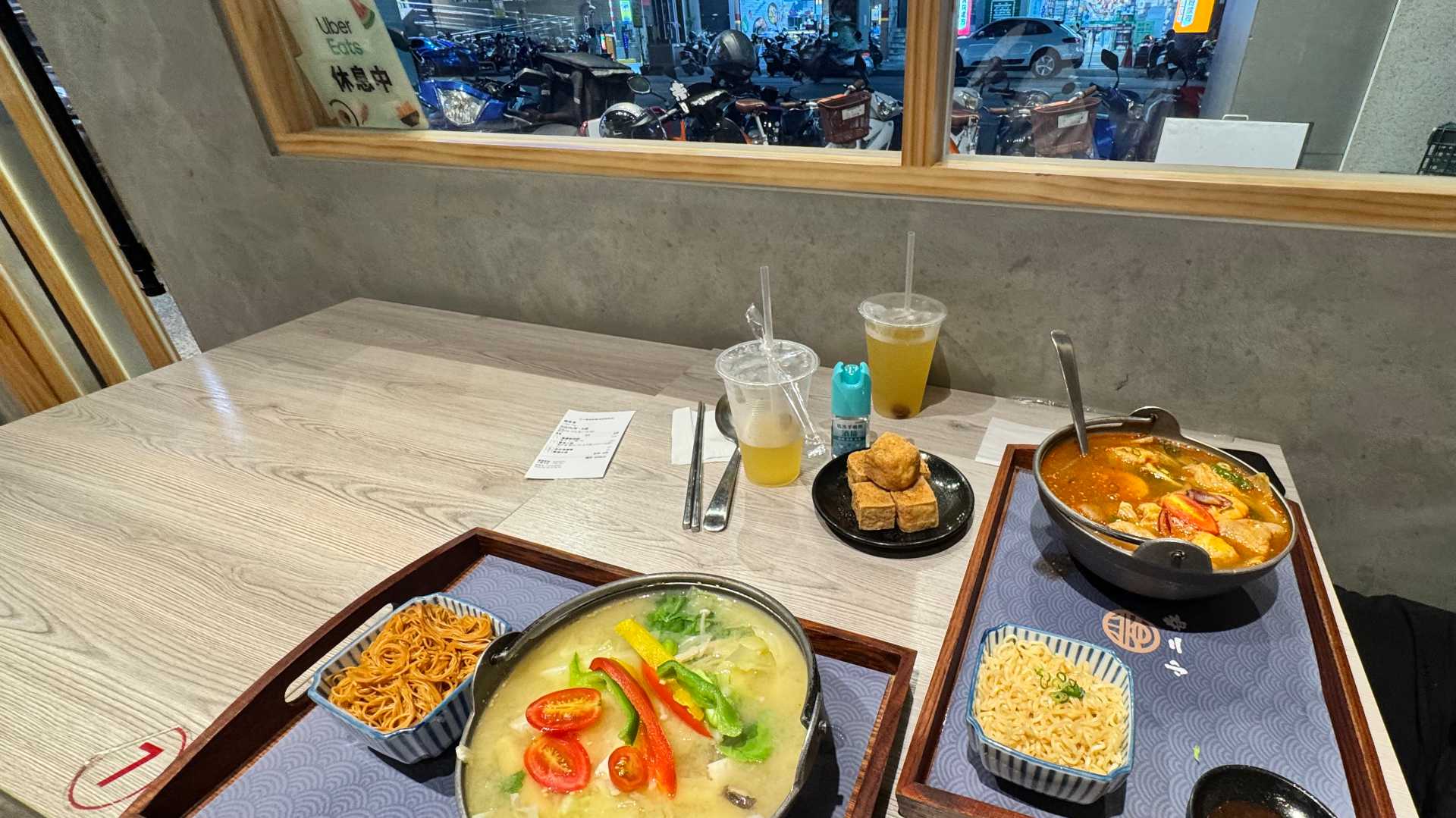 Two hotpot meals and side dishes on a table in a restaurant. Outside the window, scooters can be seen parked on the side of the street.