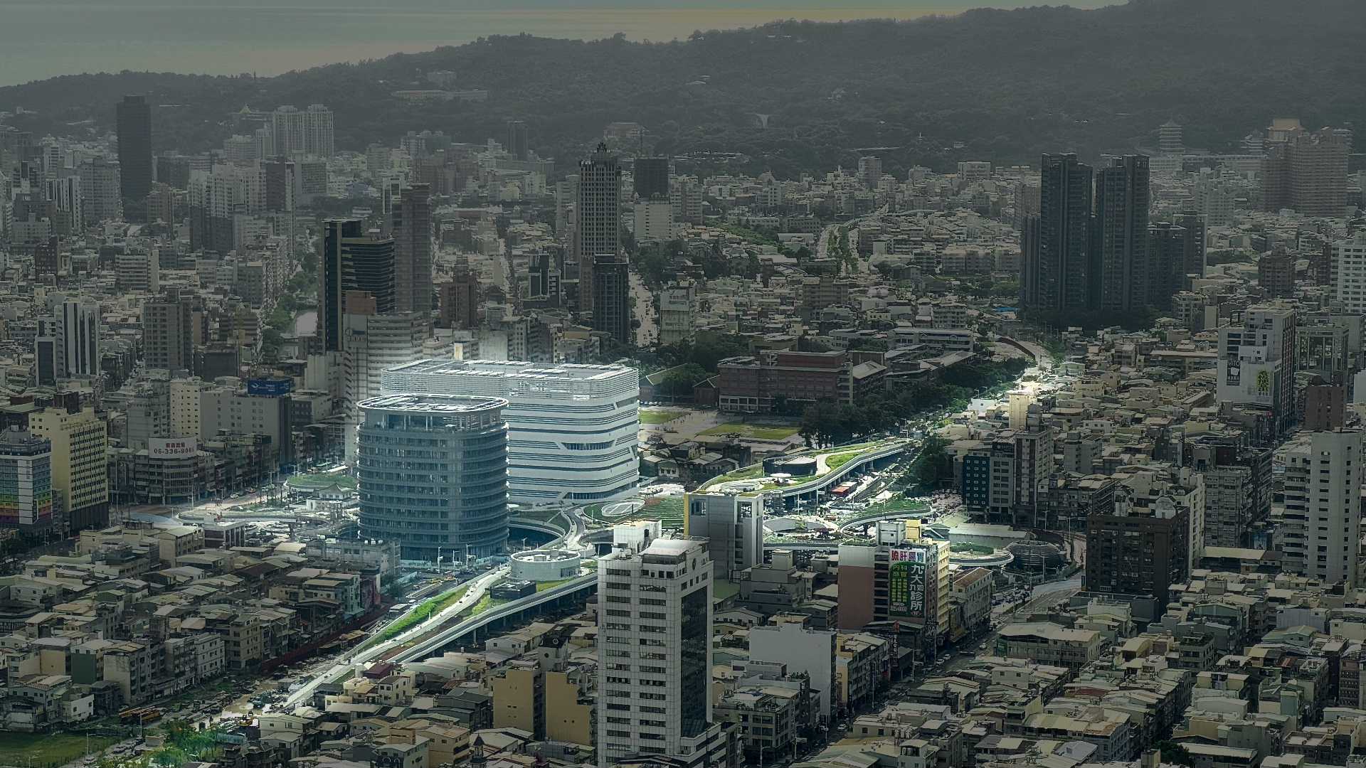 Aerial photo of Kaohsiung Main Station, under contruction, with all other areas darkened. The station comprises two towers, perhaps 10 floors each, plus a near-kilometer-long sinuous elevated grass corridor snaking through the city.