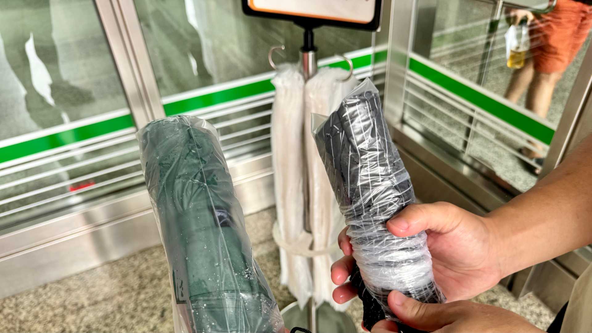 Two people holding plastic-wrapped umbrellas outside an office building entrance.