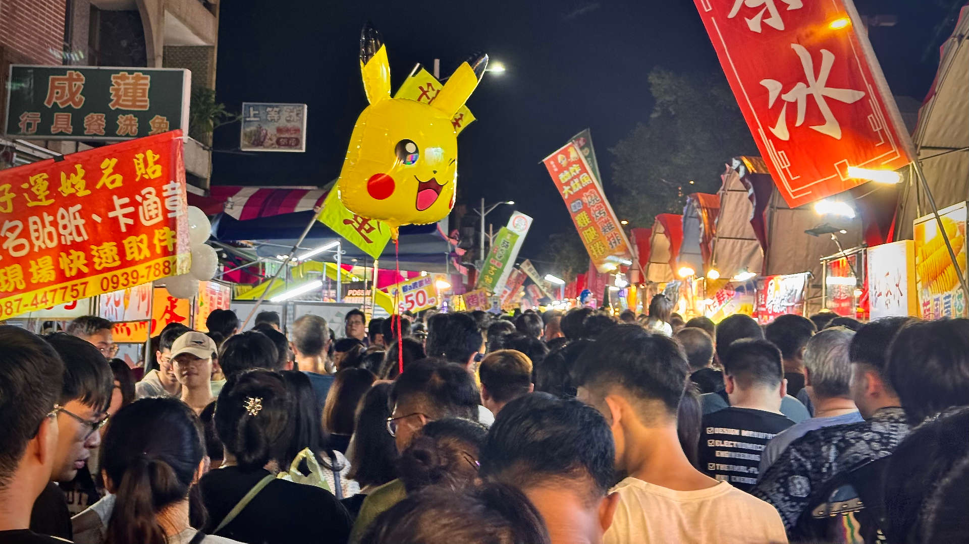 An extremely crowded night market, with a large Pikachu balloon hovering above the crowd.