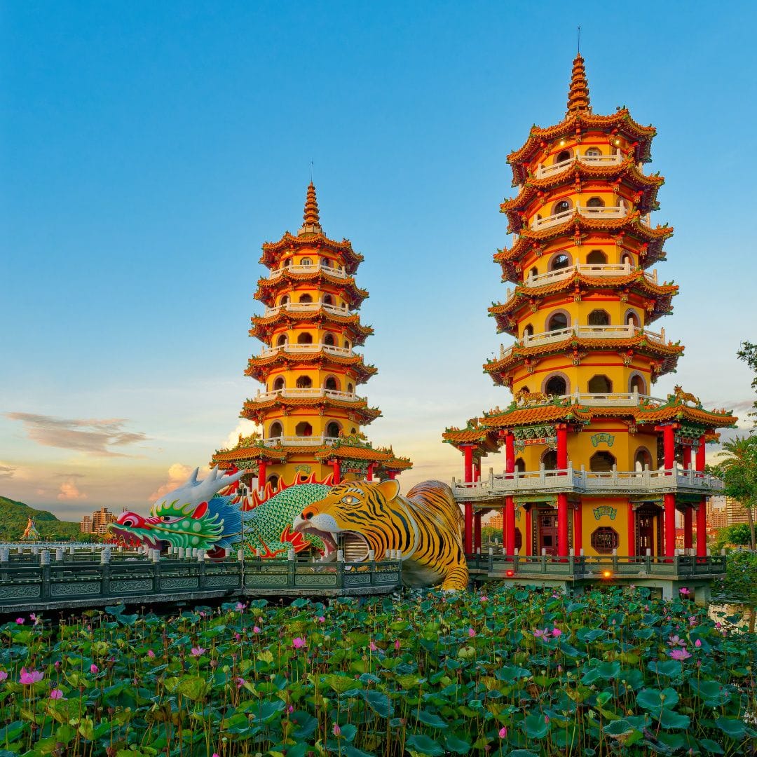 The Dragon and Tiger Pagodas at Lotus Pond, Kaohsiung, Taiwan. Pink lotus flowers are in bloom on the water. The two pagodas are eight stories tall, with an oversized dragon sculpture at the foot of one, and a tiger at the foot of the other. The mouths of these creatures are the entry and exit doors.