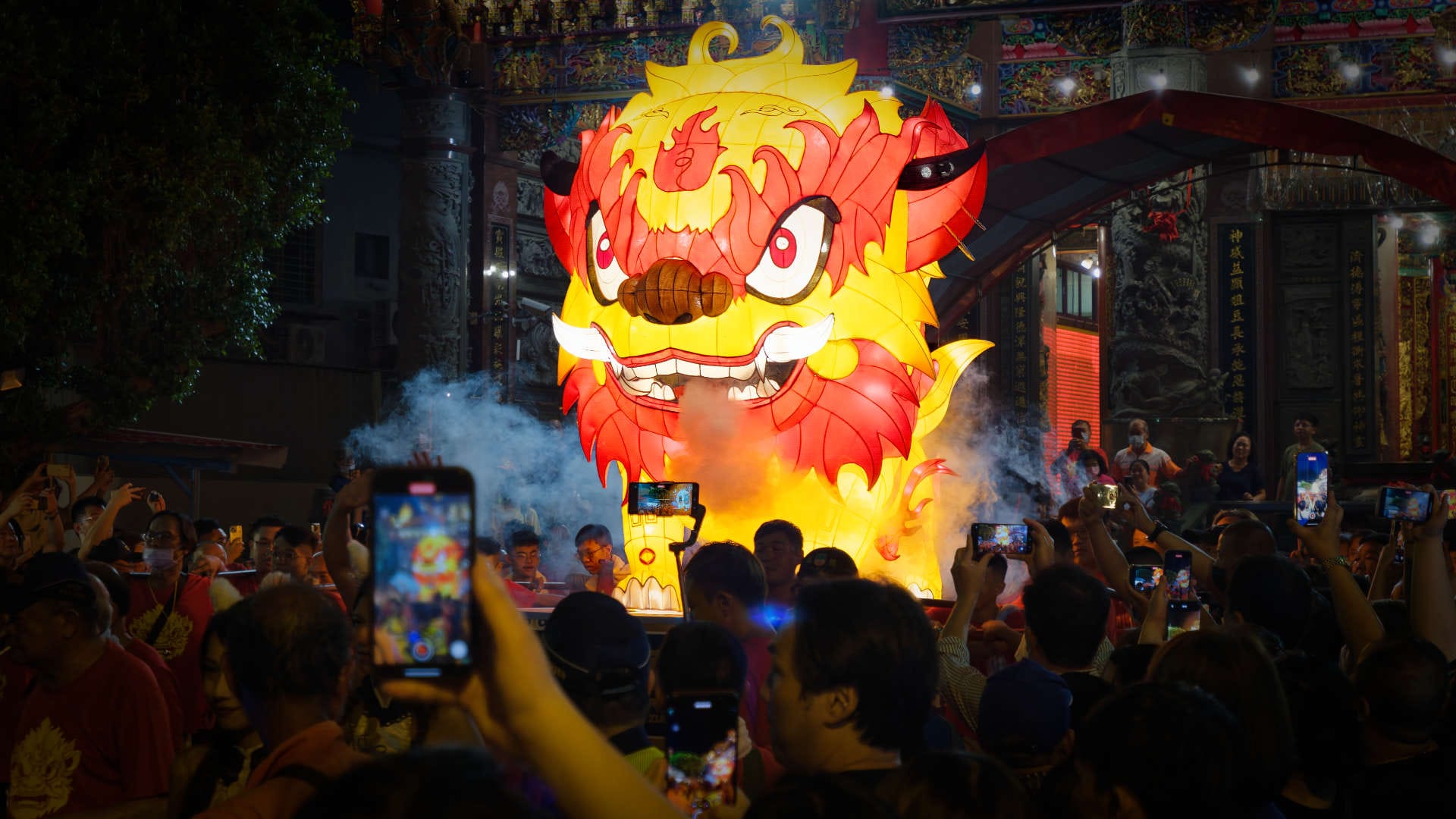 Artificial smoke coming from the mouth of a high Fire Lion lantern, held above a dense crowd of onlookers.