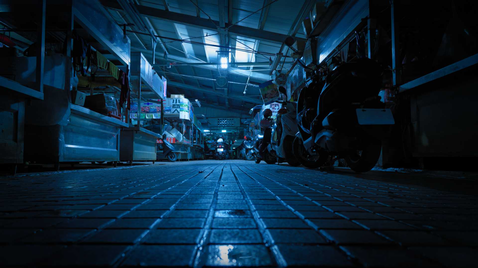 Atmospheric photo of a deserted roofed open-air market at night, with wet tiles on the floor and scooters parked alongside the empty food stalls.