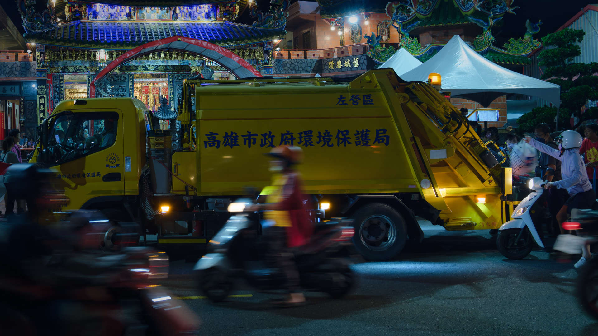 Long exposure of a Taiwanese garbage truck stopped outside a temple as people drive past on scooters. One lady is tossing a bag of garbage from her scooter into the garbage truck.