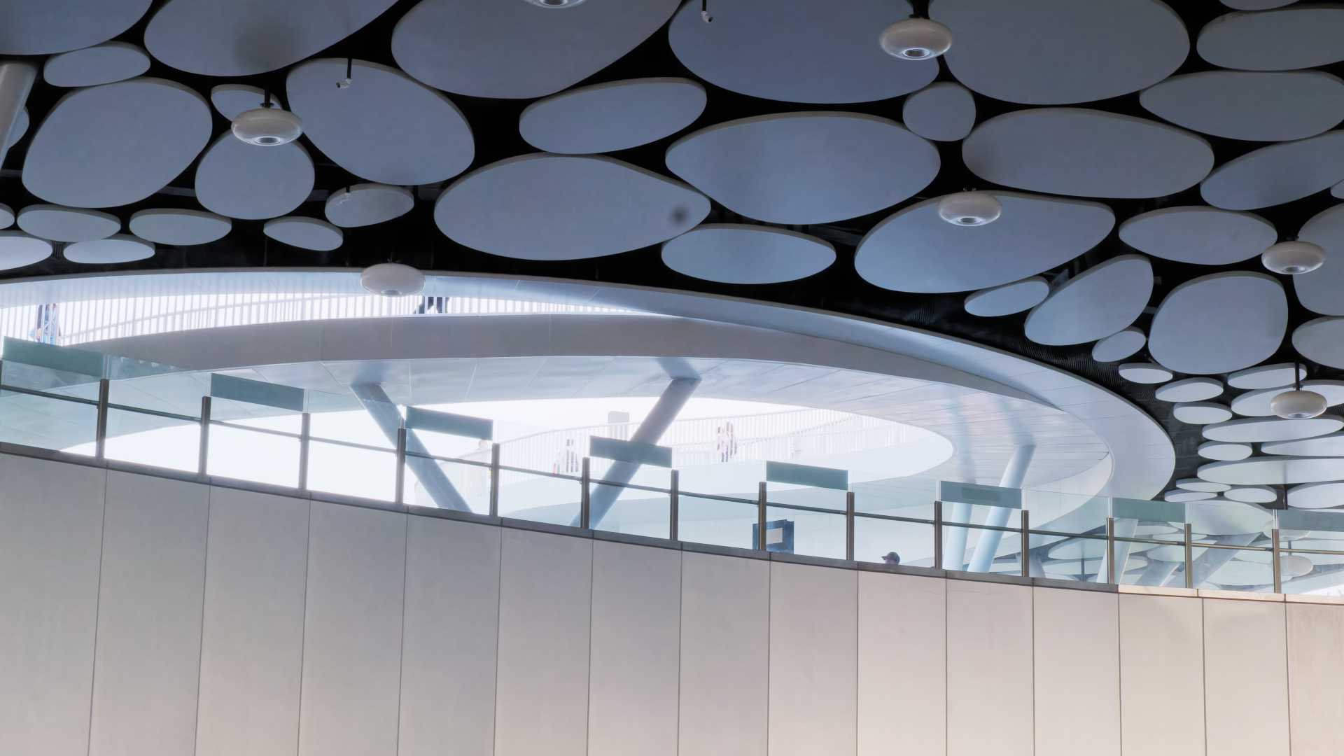 Zoomed-in view of a circular opening in the ceiling. Beyond it, people can be seen walking on a spiral ramp.