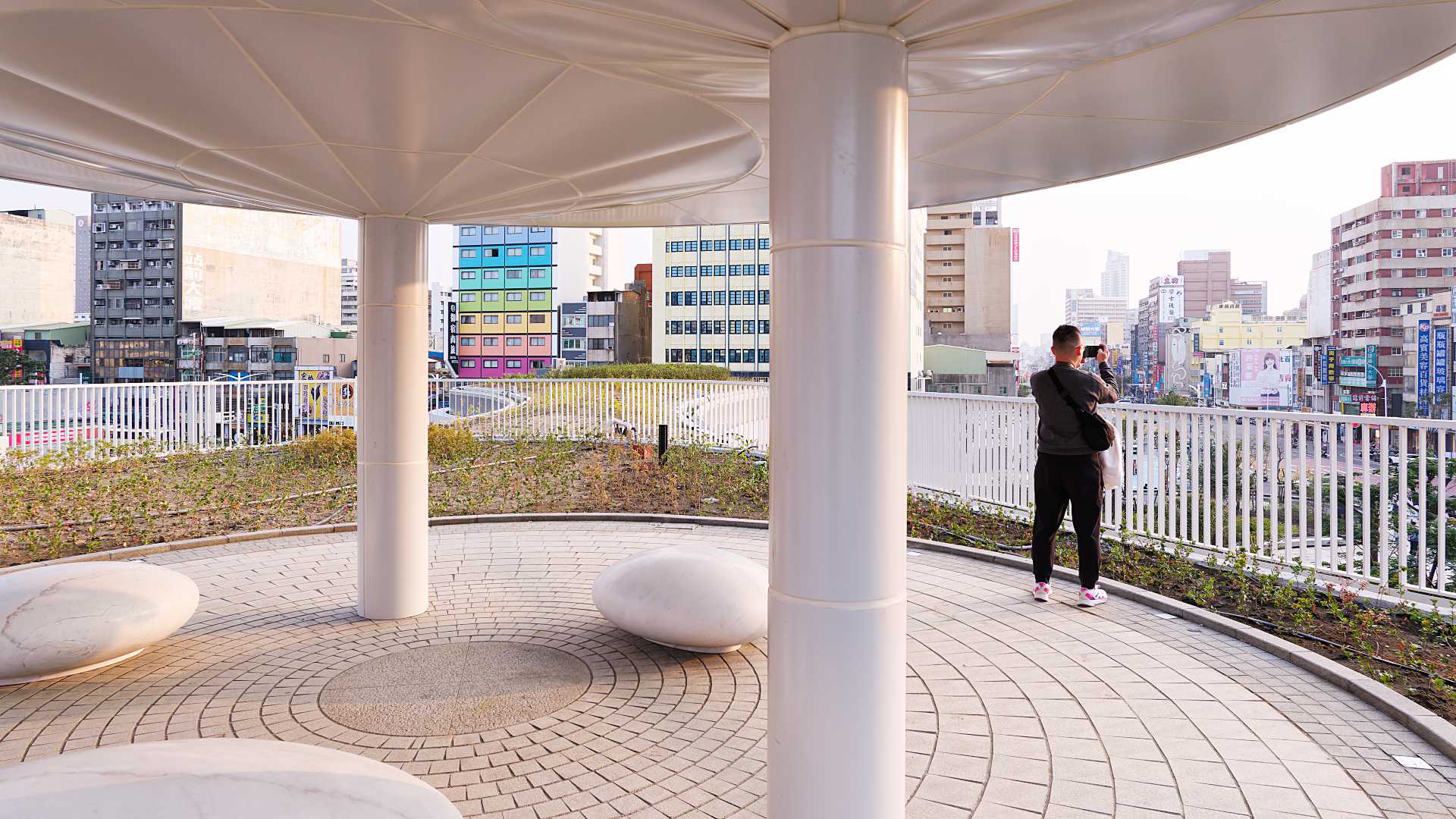 A man standing on a raised outdoor platform at Kaohsiung Station, taking a photo of the cityscape.