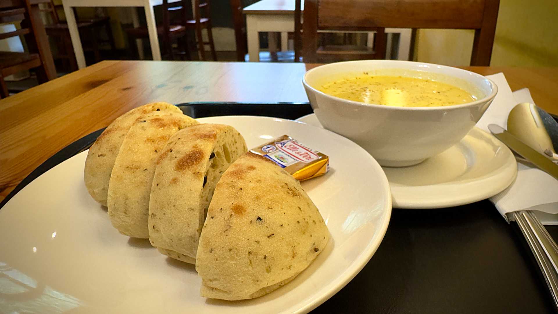 A small spicy olive bread roll slided into four pieces, on a tray alongside some butter and a bowl of vegetable soup.