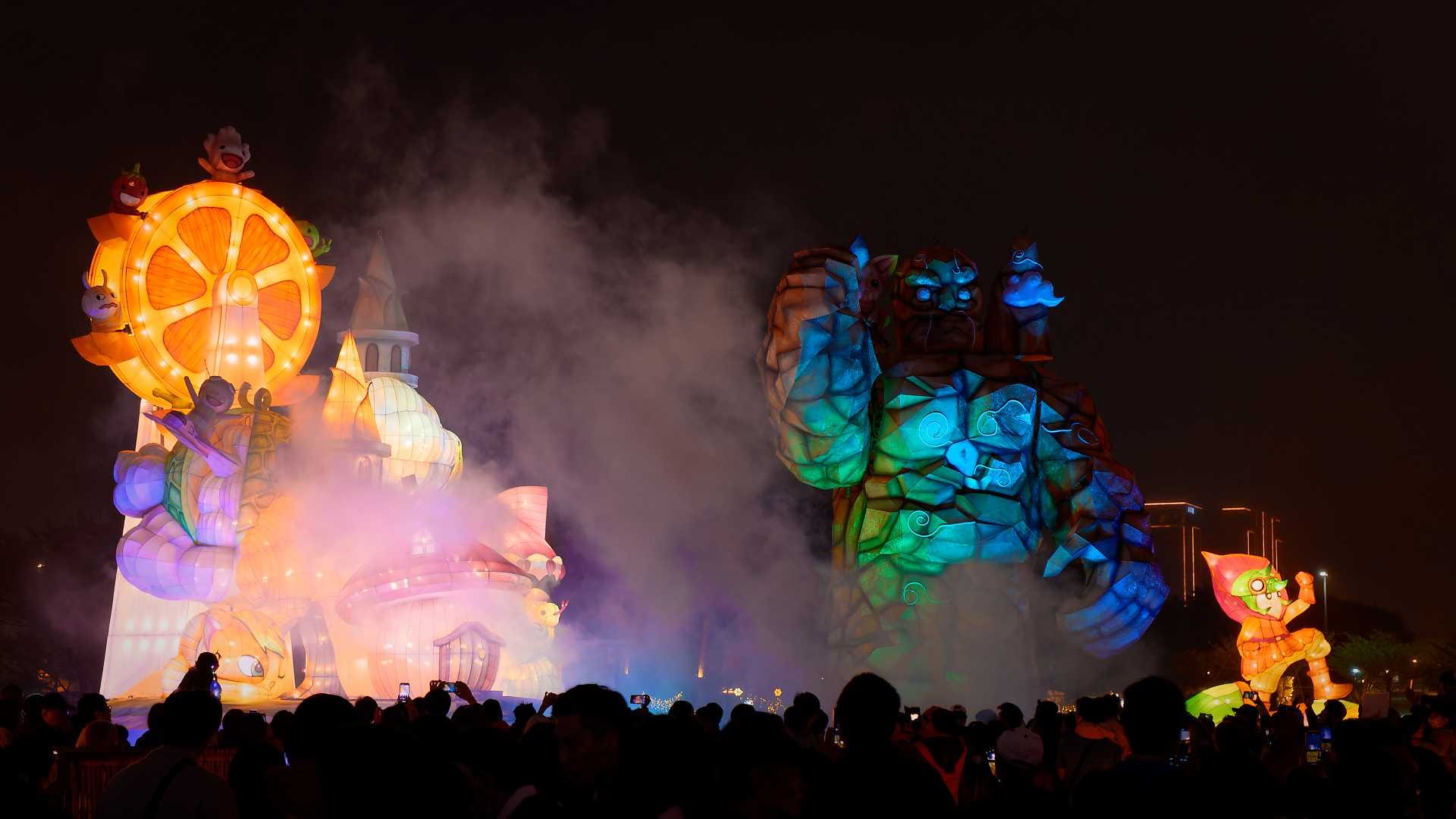 Lanterns in the shape of a cartoon ferris wheel atop a small village, and a monster made of stone. A large crowd is watching.